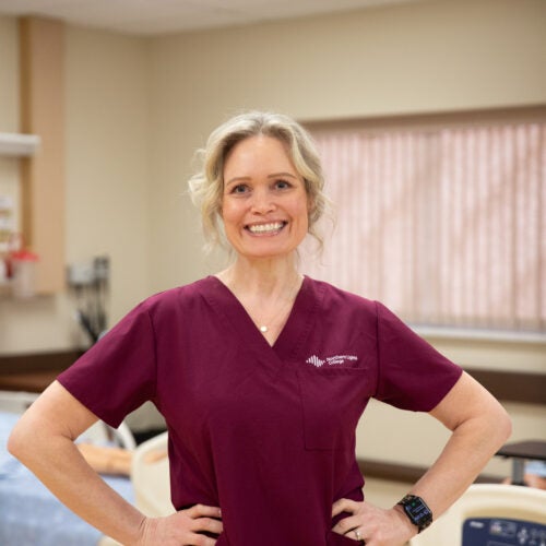April Trasy stands in the nursing lab wearing burgundy scrubs