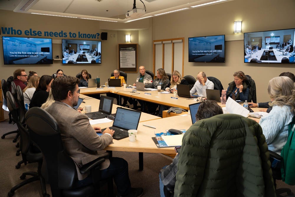 People sit around a oval-shaped table. Four screens are mounted on the walls.