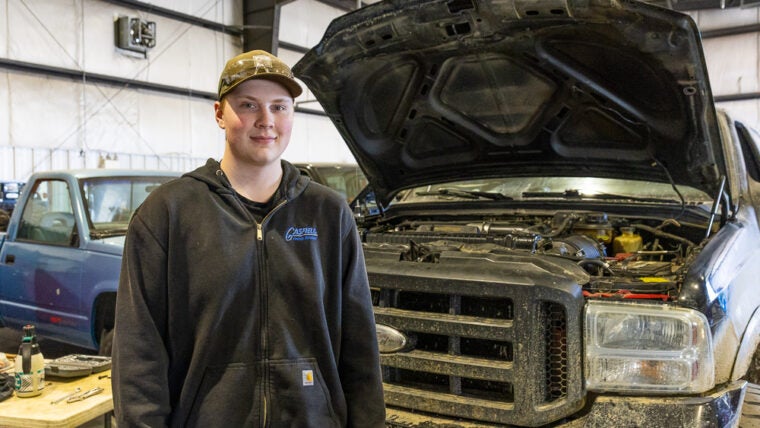A young man stands before a pickup truck with the hood raised.