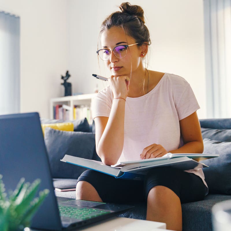 Student sitting on couch with textbook open and looking intently at computer screen